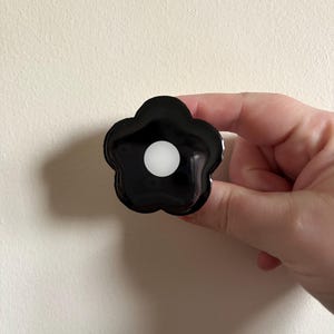 Puede incluir: Un objeto negro en forma de flor con un centro blanco. El objeto tiene un acabado brillante y se sostiene sobre un fondo neutro. El diseño floral es una forma simple y moderna.