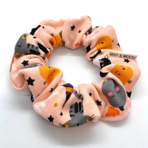 Op de afbeelding: Een roze scrunchie met een herhalend patroon van katten, sterren en manen. De scrunchie heeft de tekst "MAY & MOON" erop gedrukt.
