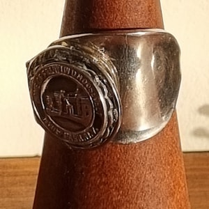 May include: A silver ring with a raised crest featuring a building and the text "University of Dallas" around the edge.