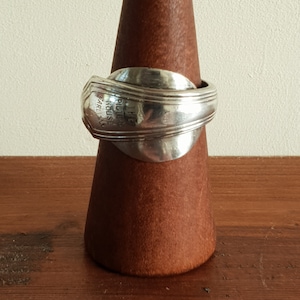 May include: A silver spoon ring with the words "The Pilgrim House" engraved on the band. The ring is displayed on a wooden ring holder.