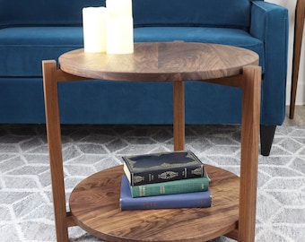 May include: A two-tiered, round wooden coffee table with a dark finish, holding books on the lower tier and candles on top. Two matching plant stands with plants are in the background, near a blue sofa.