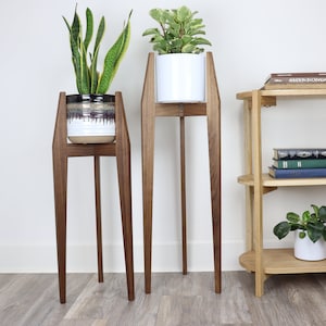 May include: Two wooden plant stands with a white ceramic planter on the taller stand and a brown and white ceramic planter on the shorter stand. The stands have a mid-century modern design with tapered legs. A wooden shelf with two shelves is in the background.