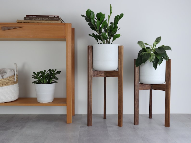 May include: Three white ceramic plant pots on wooden stands. The stands are made of dark brown wood and have a simple, modern design. The pots are all different sizes and are filled with green plants.