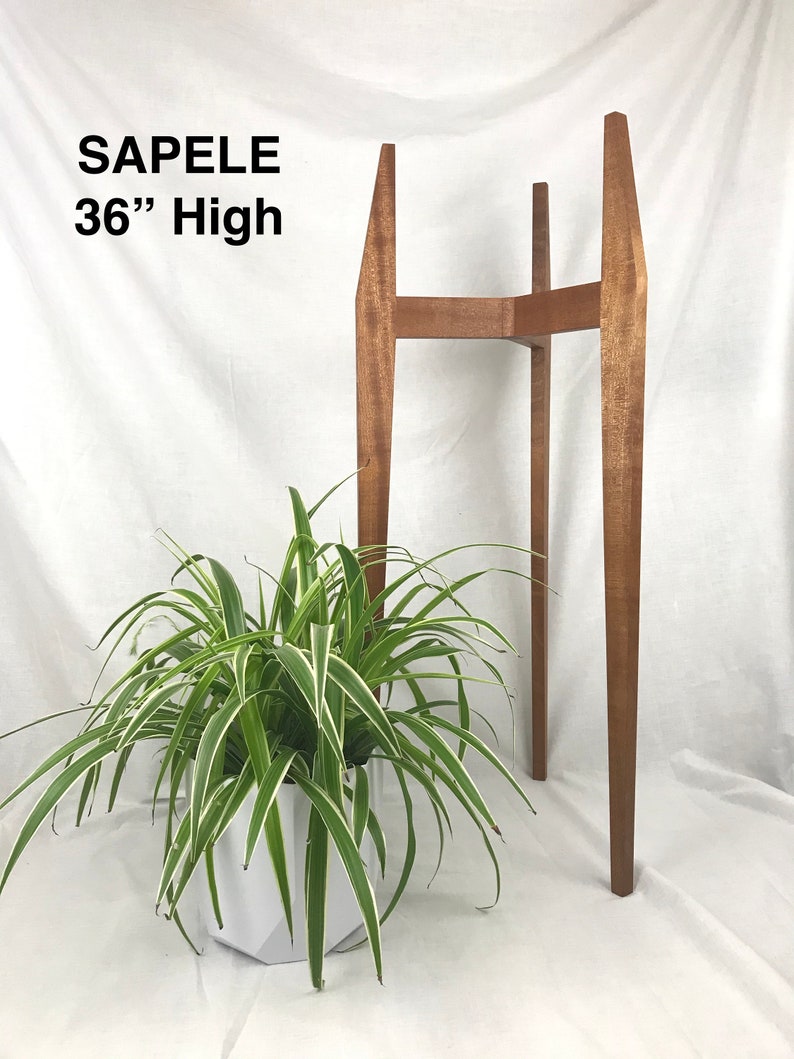 May include: A wooden plant stand made of sapele wood. The stand is 36 inches tall and has a rectangular top. The stand is shown with a green plant in a white pot.