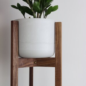 Puede incluir: Una maceta de cer&aacute;mica blanca se asienta sobre un soporte de planta de madera de nogal. El soporte tiene un dise&ntilde;o simple y moderno con cuatro patas y una base cuadrada.