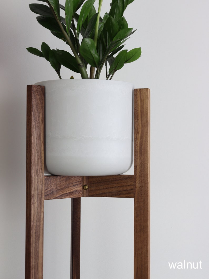 Puede incluir: Una maceta de cer&aacute;mica blanca se asienta sobre un soporte de planta de madera de nogal. El soporte tiene un dise&ntilde;o simple y moderno con cuatro patas y una base cuadrada.