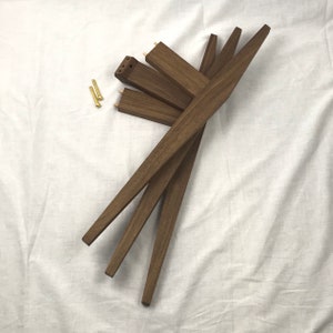 May include: Four brown wooden legs for a piece of furniture. The legs are stacked on top of each other and have a slight curve to them. Two small gold screws are visible in the background.