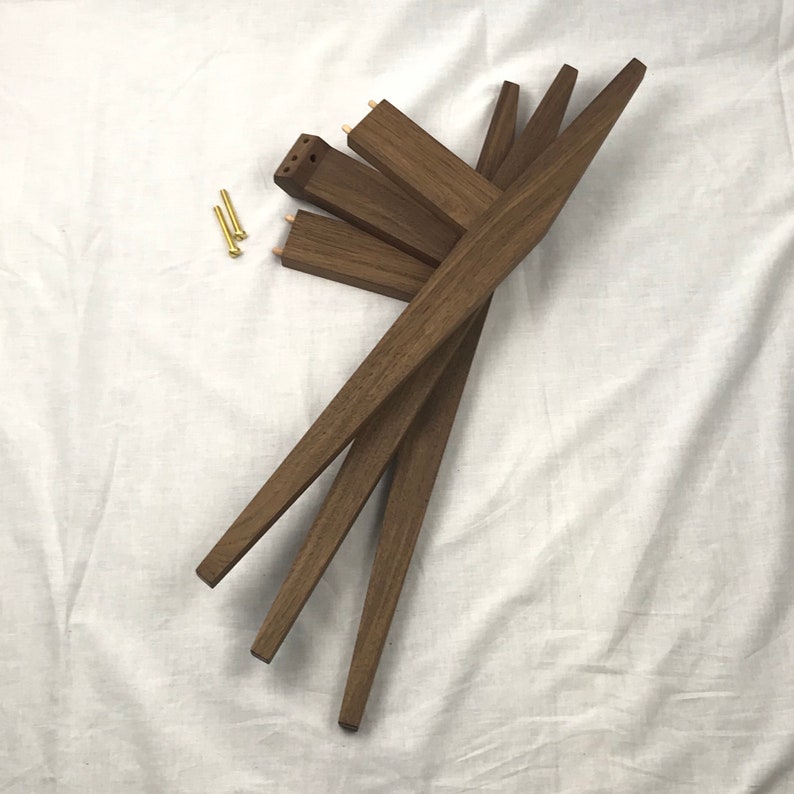 May include: Four brown wooden legs for a piece of furniture. The legs are stacked on top of each other and have a slight curve to them. Two small gold screws are visible in the background.