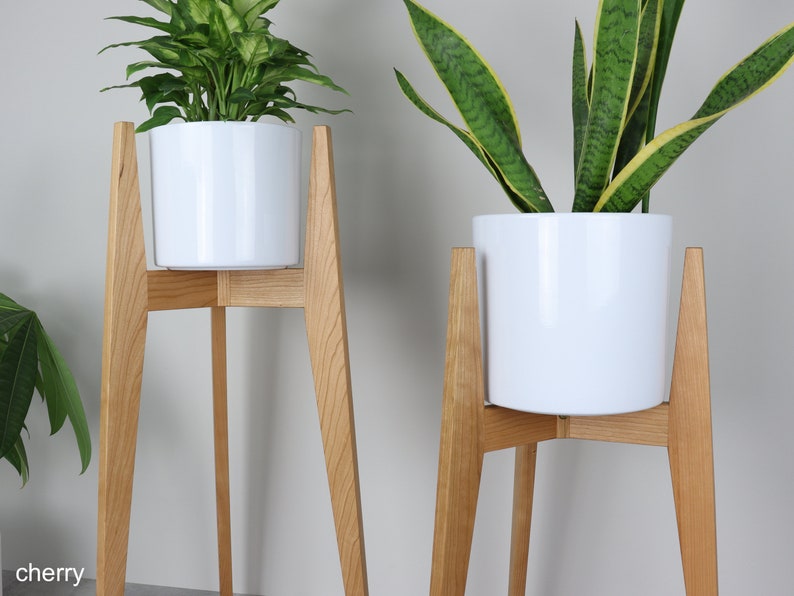 May include: Two white ceramic plant pots on wooden stands. The stands are made of cherry wood and have a mid-century modern design. The plants are green and leafy.