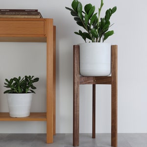 Puede incluir: Una maceta blanca con una planta verde se asienta sobre un soporte de planta de madera de nogal. El soporte tiene cuatro patas y una parte superior cuadrada. El soporte de planta est&aacute; sobre un suelo de baldosas grises.