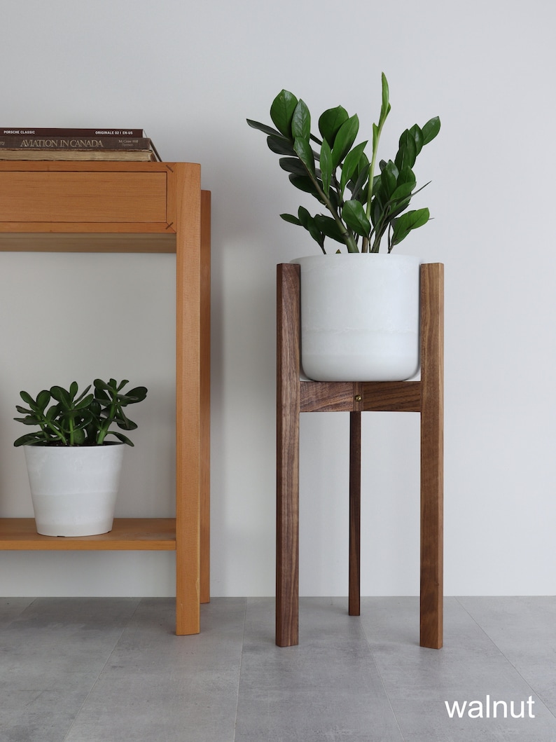 Puede incluir: Una maceta blanca con una planta verde se asienta sobre un soporte de planta de madera de nogal. El soporte tiene cuatro patas y una parte superior cuadrada. El soporte de planta est&aacute; sobre un suelo de baldosas grises.