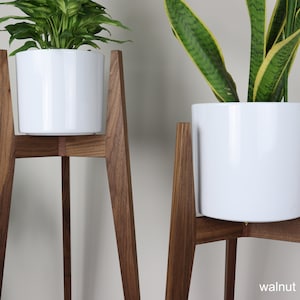 May include: Two white ceramic plant pots on wooden plant stands. The stands are made of walnut wood and have a mid-century modern design. The stands are 3-legged and have a triangular base.