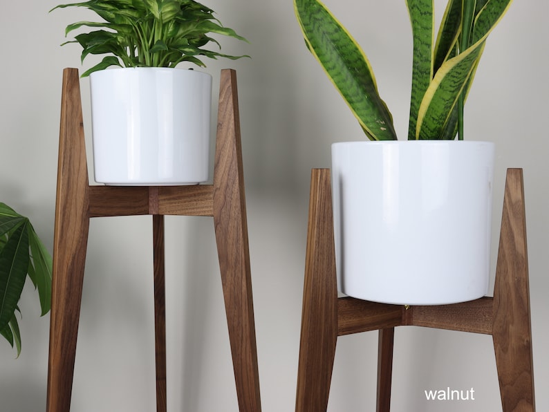 May include: Two white ceramic plant pots on wooden plant stands. The stands are made of walnut wood and have a mid-century modern design. The stands are 3-legged and have a triangular base.