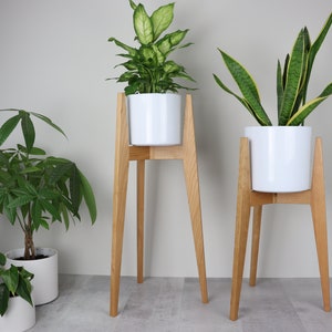 May include: Two wooden plant stands with white ceramic pots and green plants. The stands have four legs and are made of natural wood.
