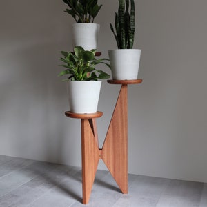 May include: A wooden plant stand with three tiers, each holding a white ceramic pot with a green plant. The stand is made of reddish-brown wood and has a triangular shape.