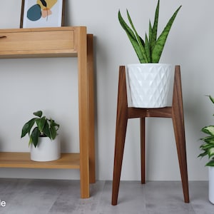 May include: A white planter with a geometric pattern sits on a wooden plant stand with four legs. The stand is made of dark wood and is about 30 inches tall. A green plant is growing in the planter. The plant stand is in front of a wooden console table with a drawer. The console table is against a white wall.