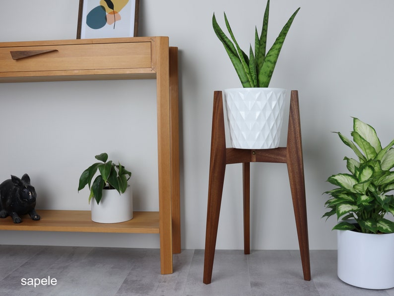 May include: A white planter with a geometric pattern sits on a wooden plant stand with four legs. The stand is made of dark wood and is about 30 inches tall. A green plant is growing in the planter. The plant stand is in front of a wooden console table with a drawer. The console table is against a white wall.