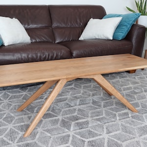 Peut inclure: Une table basse en bois clair avec un design de pieds angulaire unique. La table rectangulaire repose sur un tapis gris à motifs géométriques. Un canapé en cuir marron et des coussins sont en arrière-plan.