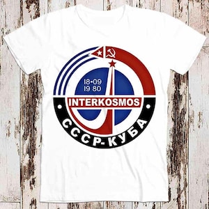 Peut inclure: T-shirt blanc avec un logo circulaire rouge, blanc et bleu. Le logo présente une étoile rouge, un drapeau rayé rouge et blanc et le texte "INTERKOSMOS CCCP-KUBA 18-09 19-80".