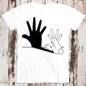 Puede incluir: Una camiseta blanca con una huella de mano negra y un diseño de sombra de conejo blanco. La huella de mano forma la silueta de un conejo con sus dedos.