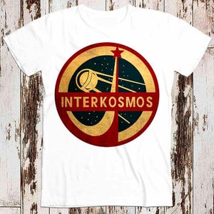 Può includere: Maglietta bianca con una grafica circolare rossa, oro e nera che presenta una navicella spaziale stilizzata e il testo "INTERKOSMOS".