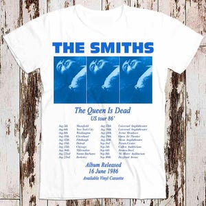 May include: White t-shirt with a blue and white graphic of the band The Smiths. The graphic includes the band's name, the album title "The Queen Is Dead", and a tour date of 1986. The album release date is also included: 16 June 1986.
