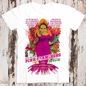 Puede incluir: Camiseta blanca con un gráfico colorido de la película "Pink Flamingos". El diseño presenta a Divine, la estrella de la película, sosteniendo una pistola, con texto y acentos vibrantes. La camiseta es un homenaje a la película de John Waters.