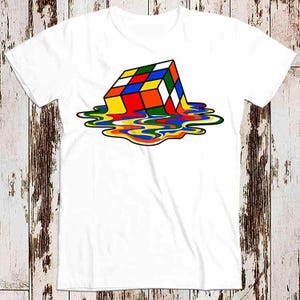 Puede incluir: Camiseta blanca con un gráfico de un Cubo de Rubik derritiéndose. El cubo se muestra con sus colores clásicos rojo, azul, amarillo, verde, naranja y blanco, con los colores goteando por la camiseta.