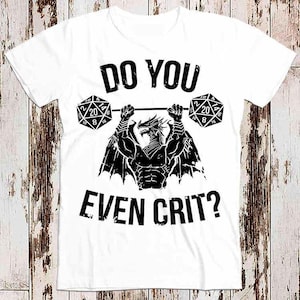 Könnte beinhalten: Weißes T-Shirt mit einem schwarzen Drachen-Design, das zwei schwarze und weiße Würfel hebt, auf denen "20" und "10" stehen. Der Text "Do you even crit?" ist unter dem Drachen aufgedruckt.