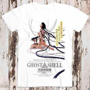 Può includere: Maglietta bianca con una grafica di una donna con fili che escono dal suo corpo. Il testo "Ghost in the Shell" è stampato sulla maglietta, insieme al testo giapponese "攻殼機動隊".