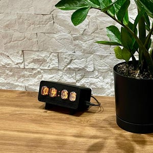 Könnte beinhalten: Eine schwarze Digitaluhr, die die Uhrzeit 09:56 anzeigt. Die Uhr hat eine rechteckige Form mit Retro-Design. Sie steht auf einer Holzoberfläche neben einer Topfpflanze mit grünen Blättern und einem weißen Lautsprecher. Die Uhr ist an eine Stromquelle angeschlossen.