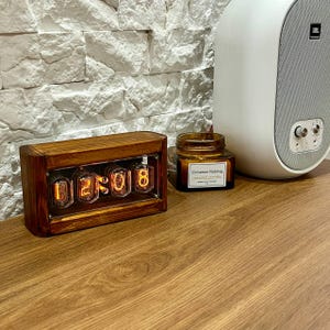Könnte beinhalten: Eine digitale Holzuhr mit der Anzeige "02:08" mit orangefarbenen, beleuchteten Ziffern. Die Uhr ist in einem rechteckigen, dunklen Holzrahmen eingefasst. Ein kleines Glas mit "Cinnamon Nutmeg" Duftkerze befindet sich neben der Uhr.