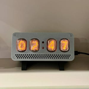 Könnte beinhalten: Eine graue Digitaluhr mit vier leuchtenden orangefarbenen Röhren, die die Uhrzeit anzeigen. Die Uhr hat eine rechteckige Form mit perforiertem Boden und wird von einem schwarzen Ständer getragen. Ein schwarzes Kabel erstreckt sich von der rechten Seite.