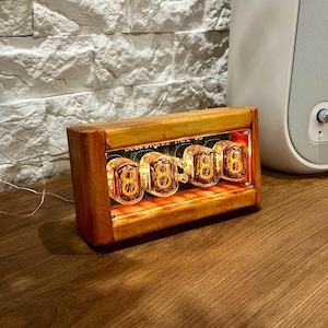 May include: A wooden clock with four glowing orange Nixie tubes displaying the time "88:88". The clock is labeled "Dekatron IN12 6S" and is sitting on a wooden surface.