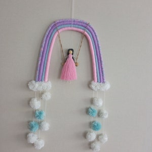 Puede incluir: Un adorno de pared de arcoíris hecho a mano con un columpio. El arcoíris presenta colores pastel de morado, rosa y azul. Una pequeña muñeca con un vestido rosa está sentada en un columpio de madera. Pompones blancos y azules cuelgan debajo.