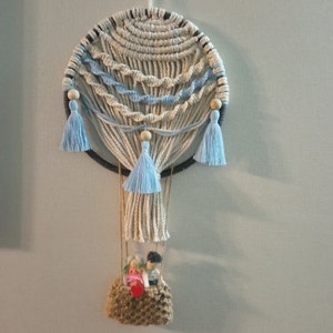 May include: A macrame hot air balloon wall hanging with blue tassels and a small woven basket with two figures inside.