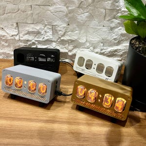 Könnte beinhalten: Eine Sammlung digitaler Uhren in verschiedenen Farben, darunter Schwarz, Weiß, Grau und Gold. Jede Uhr zeigt leuchtende orangefarbene Ziffern in einem rechteckigen Gehäuse an. Die Uhren sind auf einer Holzoberfläche angeordnet, mit einer Pflanze in einem schwarzen Topf im Hintergrund.