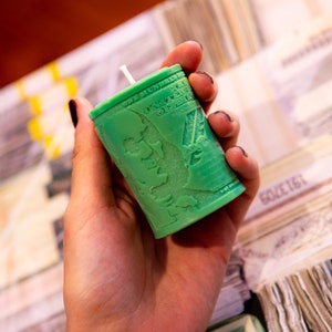 May include: A green candle shaped like a stack of money, featuring a portrait and text from a US one-hundred-dollar bill. The candle has a white wick and is held in a hand. The background shows stacks of money.