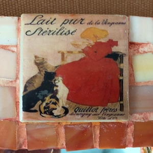 May include: A vintage tile featuring a woman in a red dress with two cats. The tile has the text "Lait pur de la Vingraine Sterilise" and "Quillot Freres Leurgy sur Vingraine Cote d'Or".