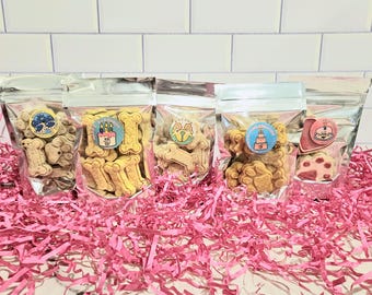 May include: Five clear plastic bags filled with dog treats. The bags are arranged in a row on a pink and white shredded paper background. Each bag has a different birthday-themed design on the treats.