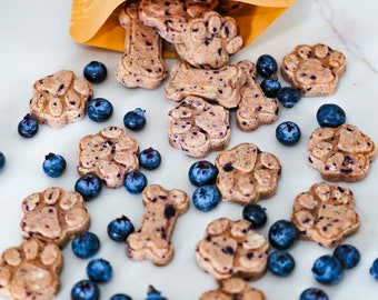 May include: A white surface with a variety of dog treats in the shapes of bones and paw prints. The treats are light brown with dark blue blueberries scattered around them. A yellow bag with the text "LOVE PAWS" is in the upper left corner of the image.