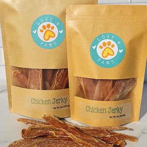 May include: Two brown stand-up pouches of Love 4 Paws Chicken Jerky with clear windows showing dried meat strips. The larger bag is 6 oz (170.1 g) and the smaller is 3 oz (85.05 g). A pile of dried chicken jerky strips sits in the foreground.