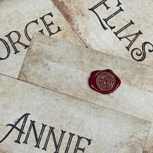 Op de afbeelding: Vier verouderd ogende enveloppen met namen in zwart gedrukt: George, Elias en Annie. Een rood waszegel met een gouden embleem staat op een envelop. De enveloppen hebben een getextureerde, antieke papieruitstraling.