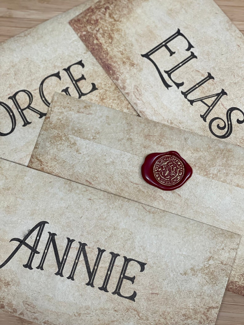 Op de afbeelding: Vier verouderd ogende enveloppen met namen in zwart gedrukt: George, Elias en Annie. Een rood waszegel met een gouden embleem staat op een envelop. De enveloppen hebben een getextureerde, antieke papieruitstraling.