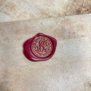 Op de afbeelding: Een bordeauxrode waszegel met gouden letters en een cirkelvormig ontwerp, met de tekst "OFFICIAL SANTA'S WORKSHOP". De zegel is bevestigd op een getextureerde, beige envelop.