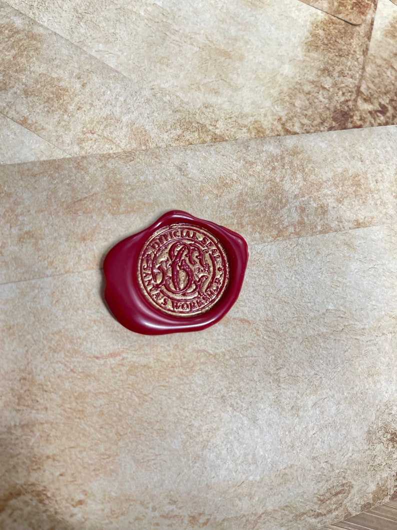 Op de afbeelding: Een bordeauxrode waszegel met gouden letters en een cirkelvormig ontwerp, met de tekst "OFFICIAL SANTA'S WORKSHOP". De zegel is bevestigd op een getextureerde, beige envelop.