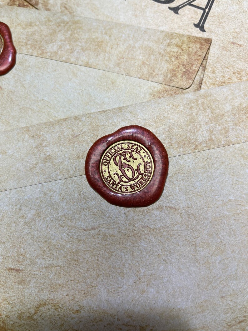 Op de afbeelding: Een close-up van een roodbruin waszegel met gouden letters met de tekst "OFFICIAL SEAL SANTA'S WORKSHOP". Het zegel bevindt zich op een cr&egrave;mekleurige envelop. De afbeelding is goed belicht en de details zijn duidelijk.
