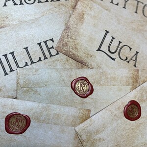Op de afbeelding: Close-up van verschillende cr&egrave;mekleurige enveloppen met namen in zwart script: Paisley, Peyton, Millie en Luca. Elke envelop is verzegeld met een rode waszegel. De zegels hebben een gouden embleem met de tekst "Official Seal, B's Workshop".
