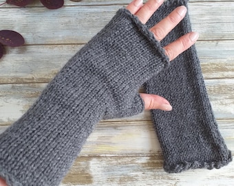 May include: A pair of charcoal gray fingerless gloves crafted from a knitted yarn material. The gloves feature a simple stockinette stitch pattern with a rolled edge at the wrist and finger openings, extending up the forearm for full coverage.