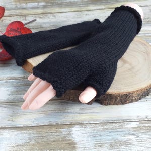 May include: A pair of black fingerless gloves made of knitted wool. The gloves are shown on a wooden surface.